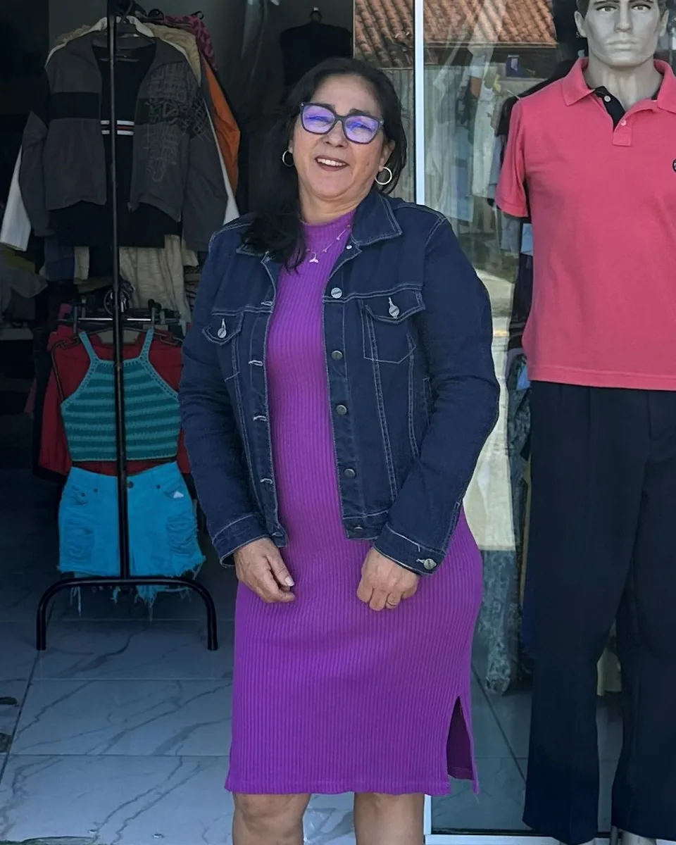 A Nenê sorrindo na porta do brechó, de vestido roxo e jaqueta jeans